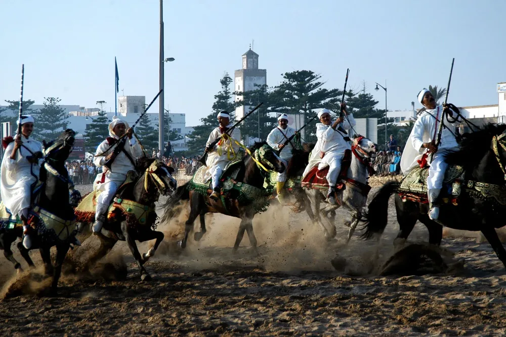 Des cavaliers en blanc chargent au galop, fusils levés, lors d’une fantasia sur la plage d’Essaouira, devant le minaret de la ville.